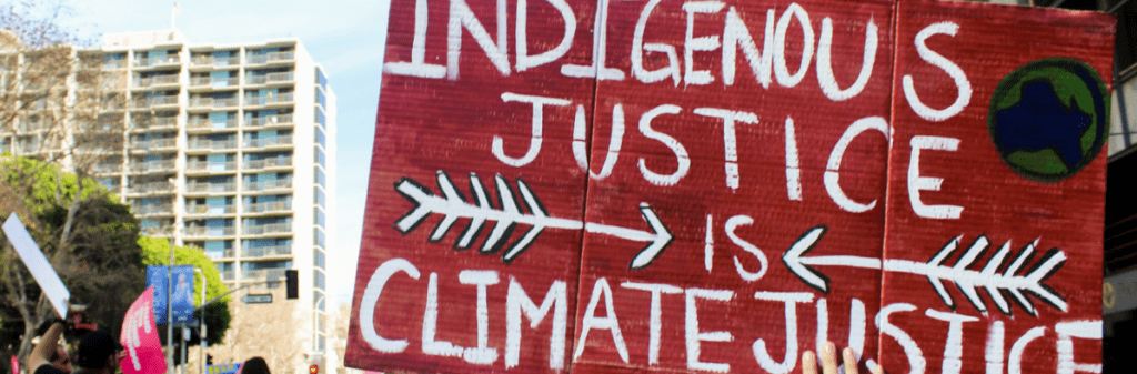 Protest sign reads "Indigenous Justice is Climate Justice" with arrows and a globe symbol.